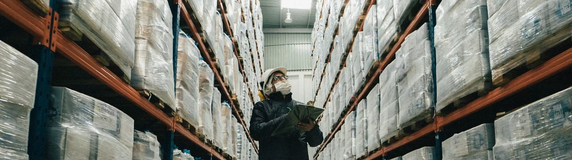 Warehouse worker checking stretch-wrapped pallets in a tall racked storage aisle